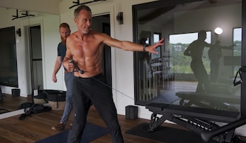 Un hombre sin camiseta realiza ejercicio con una máquina de cables mientras otro hombre con barba lo asiste en un gimnasio con espejos y una ventana