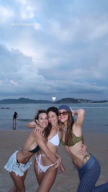 Tres mujeres sonrientes se abrazan en una playa arenosa con el mar y montañas en el fondo. El cielo está nublado