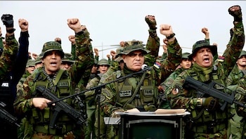 Grupo de militares con uniforme de camuflaje y armas, con varios puños levantados. Un oficial al frente habla en un podio con micrófono. Algunos parches con el texto "FANB"