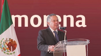 Un hombre de mediana edad con traje y corbata habla en un podio transparente. A su izquierda, se ve una bandera de México. Detrás, un muro rojo con la palabra "morena".