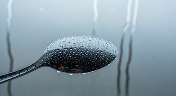Primer plano de una cuchara metálica plateada cubierta de pequeñas gotas de agua. El fondo borroso muestra una ventana empañada con marcas de humedad.