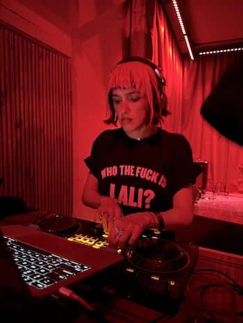 Una persona con auriculares y camiseta negra con la frase WHO THE FUCK IS LALI opera una consola de sonido bajo luz roja