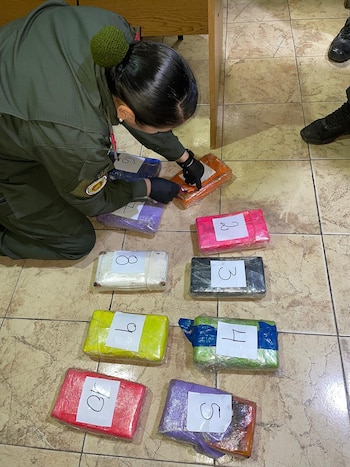 Una persona con uniforme verde y guantes negros se arrodilla sobre un suelo de baldosas claras, examinando diez paquetes rectangulares de colores variados