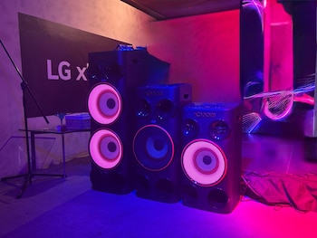 Tres altavoces negros de diversos tamaños con luces circulares magenta y azul alineados en el suelo frente a una pared