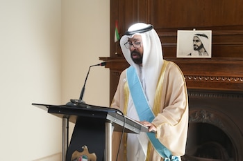 Saeed Abdulla Al Qemzi, embajador de EAU, hablando en un podio con micrófono. Viste ropa árabe tradicional. Una bandera emiratí y un retrato al fondo