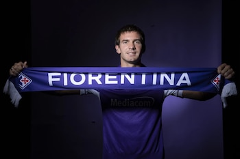 Un hombre joven sonríe y sostiene una bufanda azul y morada que dice "Fiorentina" con el logo del equipo. Viste una camiseta morada