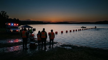 Siluetas con chalecos reflectantes a la orilla de un lago al amanecer, junto a una ambulancia encendida y una hilera de boyas en el agua.