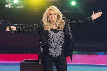 Bonnie Tyler en el escenario con cabello rubio suelto, chaqueta de lentejuelas negra y camiseta brillante, sosteniendo un micrófono con un brazo extendido