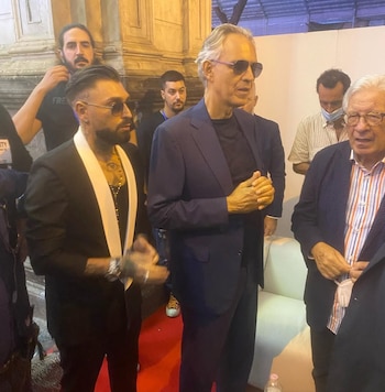 Tres hombres en atuendo formal posan sobre una alfombra roja. Un hombre con traje azul y gafas de sol, otro con traje negro de solapas blancas y el tercero con camisa a rayas