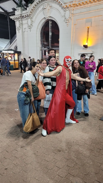 Un luchador de Titanes en el Ring con traje rojo, capa y máscara blanca posa con cinco jóvenes sonrientes frente a un edificio blanco ornamentado. Otros asistentes al fondo
