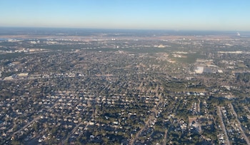 Fotografía aérea de un paisaje urbano con viviendas, calles, y una gran cantidad de árboles. Al fondo, se observa una zona con edificios y un cuerpo de agua