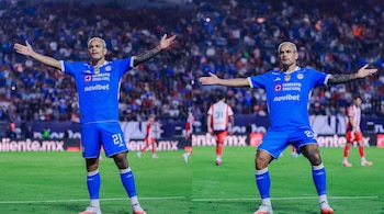Dos futbolistas con uniforme azul número 21 en un campo de fútbol, con las manos abiertas, un estadio con público en las gradas y vallas publicitarias.