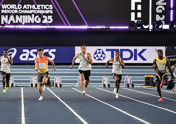 Cinco corredores disputan una carrera en una pista de atletismo azul bajo techo frente a pantallas digitales
