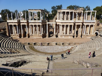 Vista panorámica del Teatro Romano de Mérida con sus gradas semicirculares de piedra, el escenario con columnas y estatuas, y personas caminando bajo un cielo azul claro