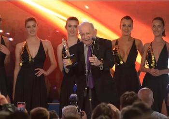 Un hombre en traje sostiene un trofeo dorado frente a un micrófono en un escenario junto a cuatro mujeres vestidas de negro