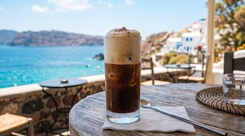 Vaso alto de capuchino frío con espuma sobre una mesa de madera, junto a una cuchara, con el mar y viviendas blancas al fondo.