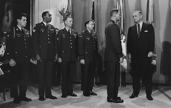 Cinco hombres de pie, cuatro con uniforme militar y uno con traje oscuro, frente a banderas y un candelabro. Es una imagen en blanco y negro
