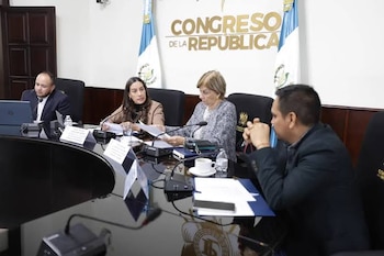 Personas en vestimenta formal sentadas ante mesas con manteles blancos, computadoras portátiles y documentos en una sala con retratos