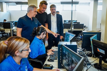 Una mujer con auriculares trabaja en un teclado de computadora, mientras tres hombres la observan y uno señala la pantalla en una sala de informática con computadoras