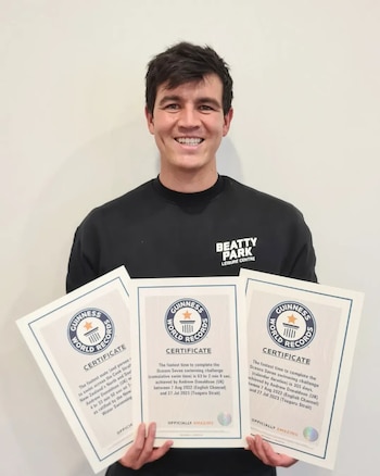 Un hombre joven y sonriente, Andy Donaldson, con un jersey negro, sostiene tres certificados Guinness World Records contra una pared blanca