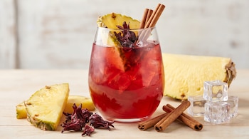 Un vaso grande de agua de piña, jamaica y canela con hielo y rodajas de piña. Alrededor, flores de jamaica, ramas de canela y cubos de hielo en madera clara.