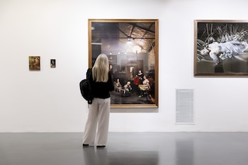 Mujer de espaldas, con cabello rubio y ropa clara, frente a una gran pintura de arte moderno enmarcada en una galería de arte con paredes blancas y suelo gris