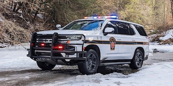 Vehículo SUV policial blanco con balizas azules y rojas en el techo, defensas delanteras negras, sobre una superficie cubierta de nieve y hielo, con árboles