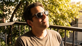 Primer plano de un hombre con gafas de sol oscuras, mirando hacia arriba bajo la luz del sol. El fondo muestra follaje borroso y una barandilla oscura.