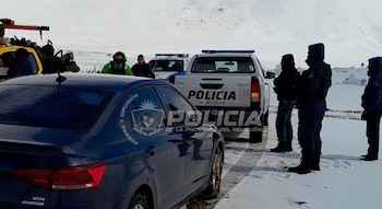 Un automóvil oscuro con cadenas en sus ruedas y una camioneta policial están sobre una carretera rodeada de montañas nevadas