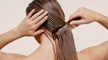 Vista trasera de una persona peinando su cabello castaño y mojado con un peine de carey mientras aplica un producto cremoso blanco.