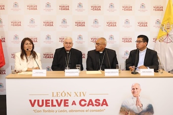 Cuatro personas sentadas ante micrófonos en una mesa frente a un panel con inscripciones y una bandera vaticana