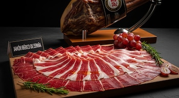 Plato de jamón ibérico de España finamente cortado, dispuesto en forma de abanico sobre una tabla de madera, con una pata de jamón en jamonero, uvas y romero.