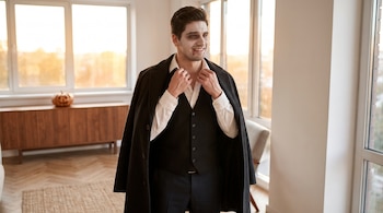 Hombre con maquillaje de vampiro, camisa blanca, chaleco y abrigo sobre los hombros, frente a un ventanal con una calabaza de fondo