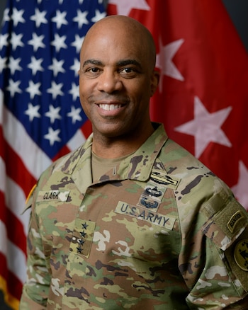 Retrato de un militar con uniforme camuflado frente a la bandera de Estados Unidos y una bandera roja con estrellas blancas