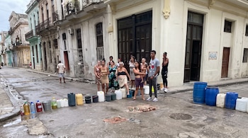 Personas de pie en una calle junto a baldes, bidones y barriles de plástico cerca de una pared con la palabra AGUA.