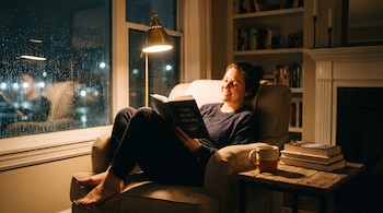 Mujer sentada en un sillón lee el libro "The Quiet Hours" junto a una ventana con lluvia. Se ve una lámpara, una mesa auxiliar con una taza y libros.