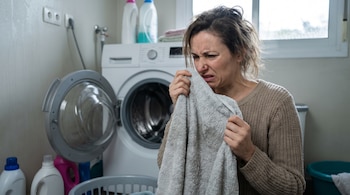 Mujer con suéter beige sujeta toalla gris cerca de la nariz con expresión de disgusto. En un cuarto de lavandería, hay una lavadora, cestos de ropa y botellas de detergente.