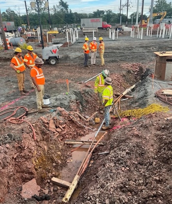 Varios trabajadores de la construcción con cascos y chalecos reflectantes observan zanjas y cables expuestos en una obra al aire libre