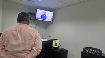 Vista posterior de un hombre con camisa frente a dos monitores que muestran a una mujer vestida con un uniforme azul.