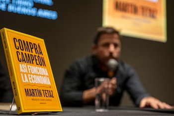 Primer plano de un libro amarillo titulado "Compra Campeón" de Martín Tetaz; en segundo plano, un hombre desenfocado habla por un micrófono