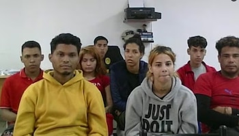 Un grupo de hombres y mujeres jóvenes, incluyendo una mujer en sudadera gris al frente, sentados en fila en una habitación blanca