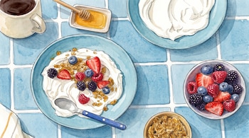 Vista superior de un tazón de yogur con granola y frutos rojos, una taza de café, un recipiente con miel, y cuencos adicionales de bayas y granola sobre azulejos azules.