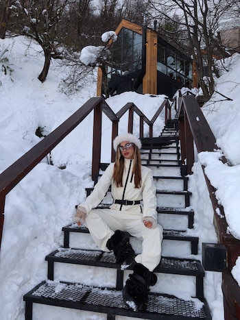 Julieta Poggio, sentada sobre los escalones nevados de la villa, con mameluco blanco y botas negras de nieve