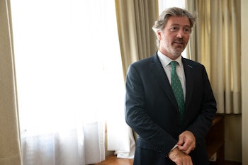 Un hombre de mediana edad con cabello gris y barba, vistiendo traje oscuro, camisa blanca y corbata verde, se ajusta el puño de la camisa en una sala bien iluminada