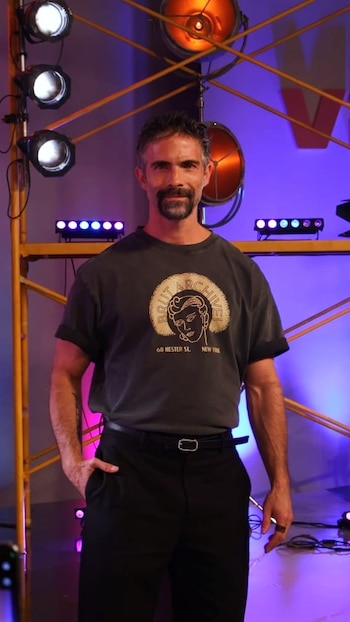 Hombre con camiseta gris y pantalón negro parado en un estudio con luces escénicas, andamios amarillos y fondo de color morado