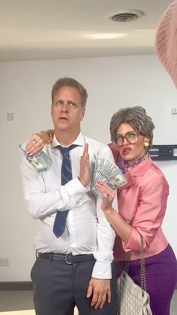 Un hombre con camisa blanca y corbata junto a una mujer con peluca gris y gafas grandes sostienen billetes de dólares estadounidenses