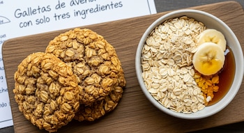 Primer plano de tres galletas de avena hechas con tres ingredientes, junto a un cuenco con avena cruda, rodajas de plátano y sirope sobre una tabla de madera.
