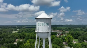 Una torre de agua blanca con el texto De Soto en letras verdes, rodeada de árboles y bajo un cielo azul con nubes