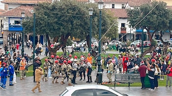 Multitud en una plaza, incluyendo personas con cascos rojos y uniformes militares que transportan una camilla. Hay árboles, farolas y edificios coloniales