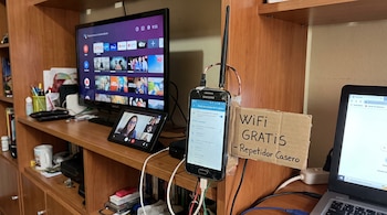 Teléfono con pantalla fisurada conectado a una antena y un cartel de cartón, junto a un televisor, una tableta y una computadora.